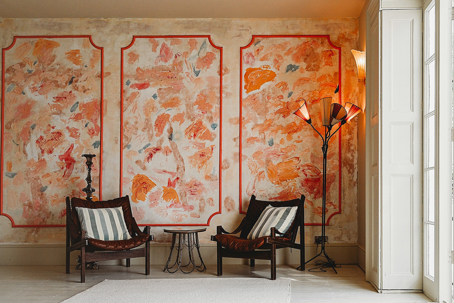 Blush pink wall with paint brush strokes over top. Colours are bright orange, muted blue and red. Two chairs and an art deco lamp sit in front.